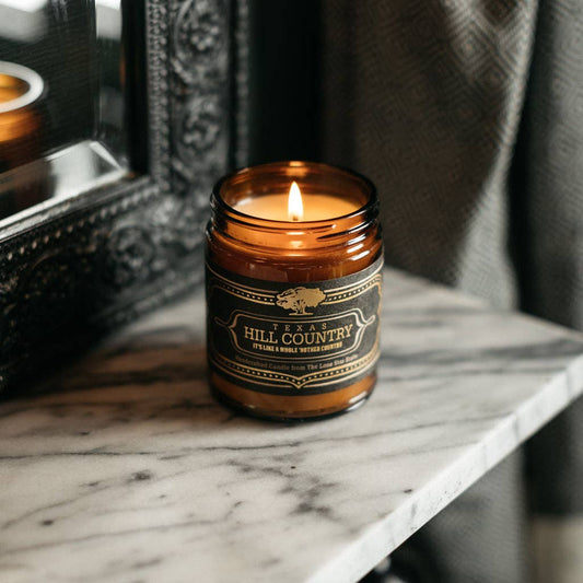 Candle in an amber jar with a label on a marble surface, with a mirror and dark fabric in the background.