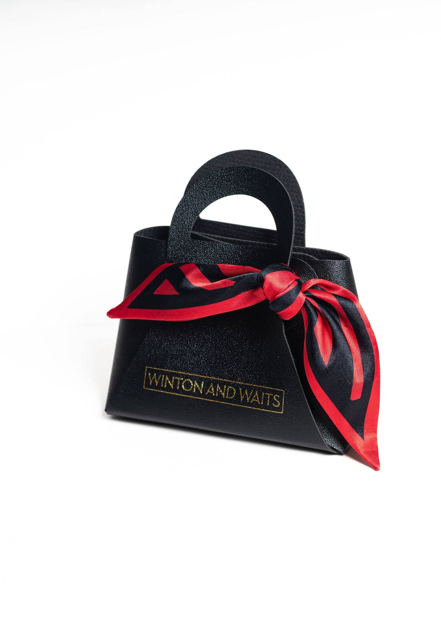 A black mini handbag with a top handle, featuring a red and black patterned silk scarf tied in a bow around the middle and gold foil branding that reads 'WINTON AND WAITS'.