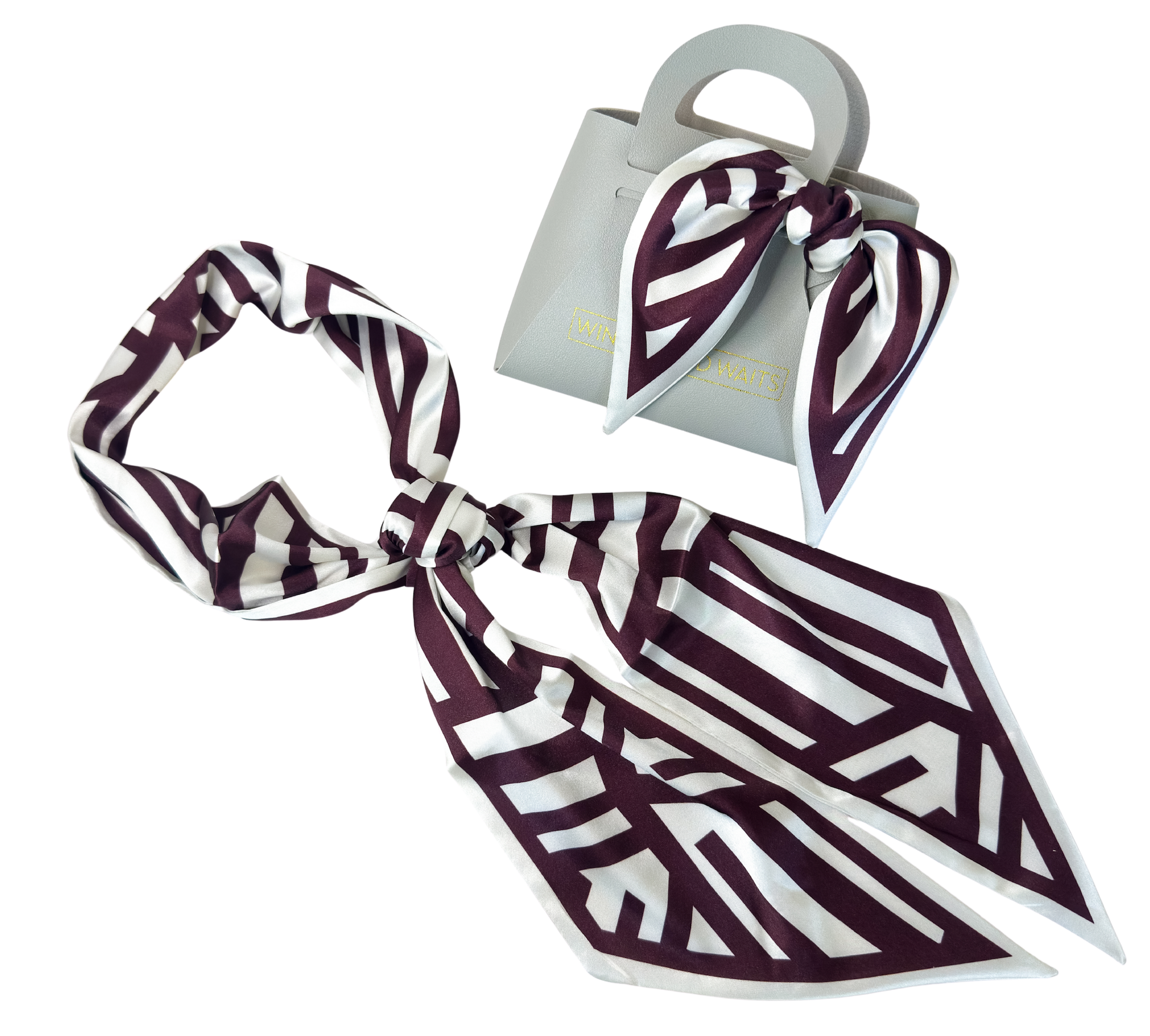 A silk scarf with a geometric art deco pattern in deep burgundy and white, shown tied in a knot next to a small light gray handbag with a matching scarf.
