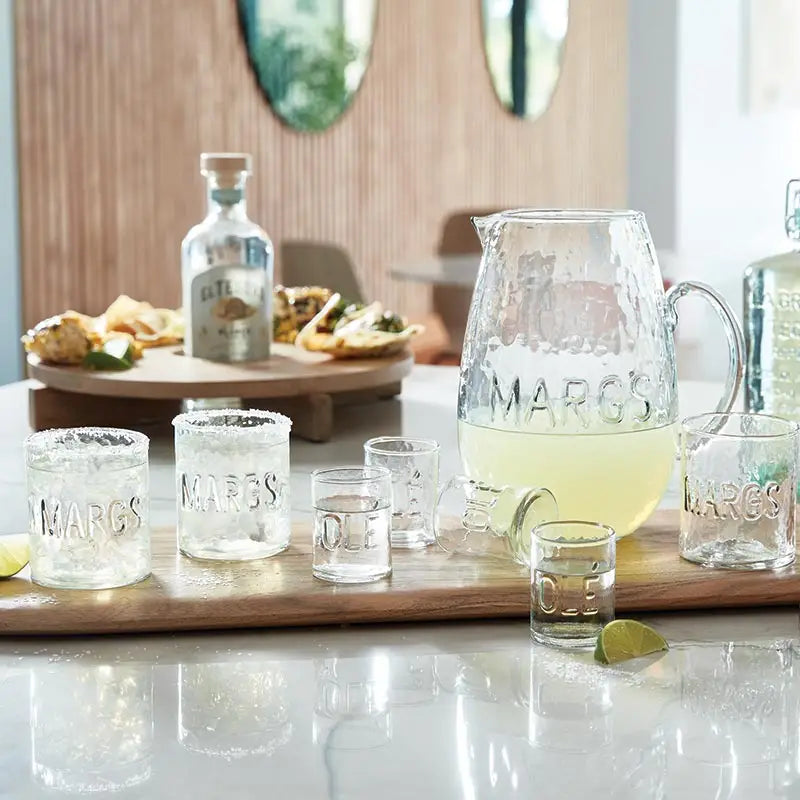 Margarita glasses and pitcher on a table with snacks and bottles in the background