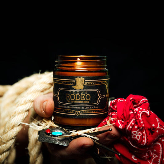 Hand holding a 'Texas Rodeo' candle with a red bandana and rope in the background