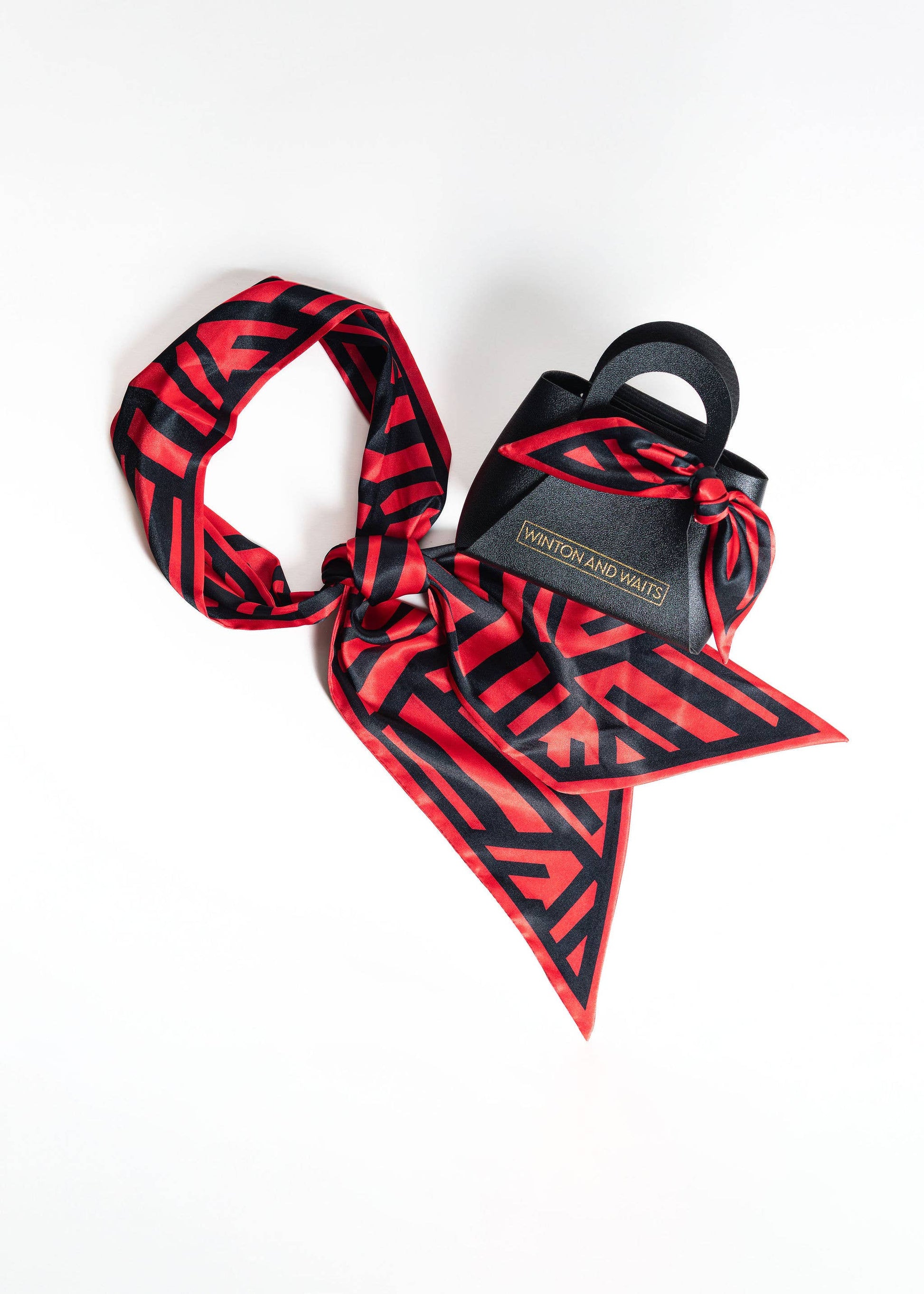 A black and red geometric patterned silk scarf styled with a small black leather handbag featuring a matching red and black patterned ribbon tie, displayed on a clean white background.