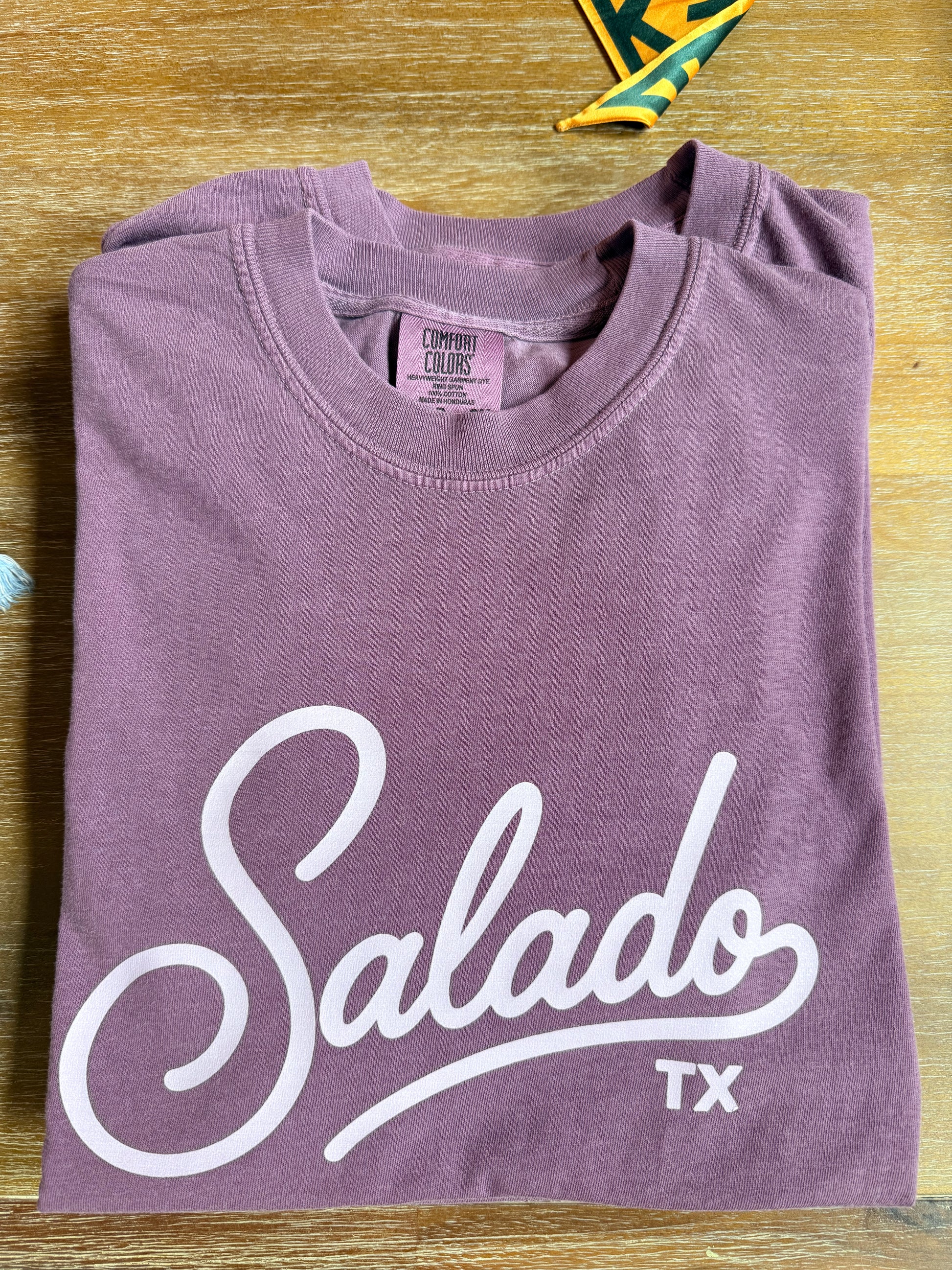 A folded heathered mauve-purple t-shirt with 'Salado TX' printed in white script across the front, laid flat on a light wood surface.