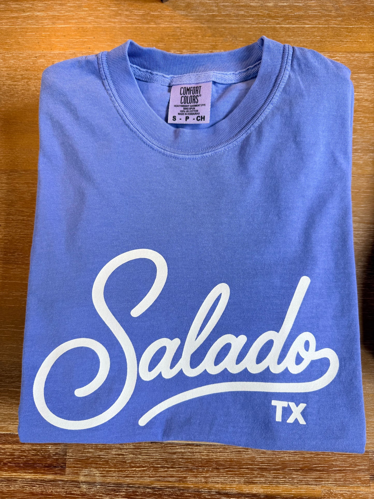 A folded blue t-shirt with a white 'Salado TX' graphic printed on the front, laid flat on a wooden surface. The neck label reads Comfort Colors.