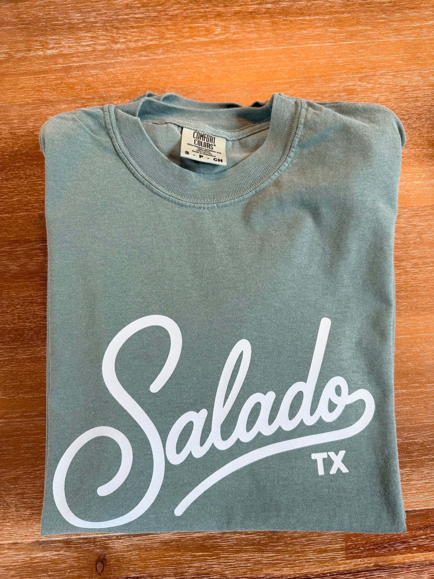 A folded sage green t-shirt with a white graphic print reading 'Salado TX' in cursive script, laid flat on a wooden surface.