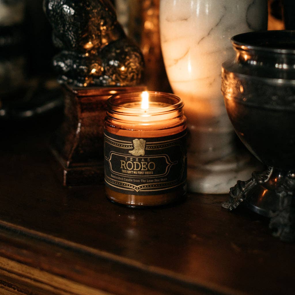 Candle in a jar labeled 'Rodeo' on a wooden surface with decorative items in the background.