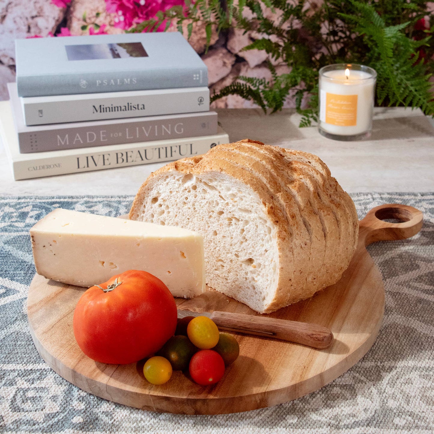 Loaf of bread, cheese, and tomatoes on a wooden board with books and a candle in the background.