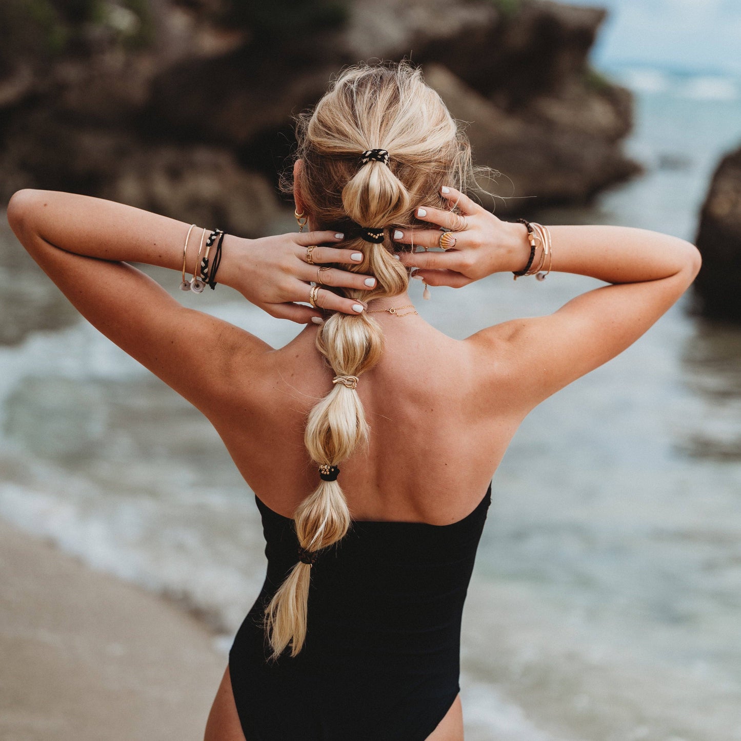 Woman in a black swimsuit with braided hair standing on a beach with rocky cliffs and ocean in the background.