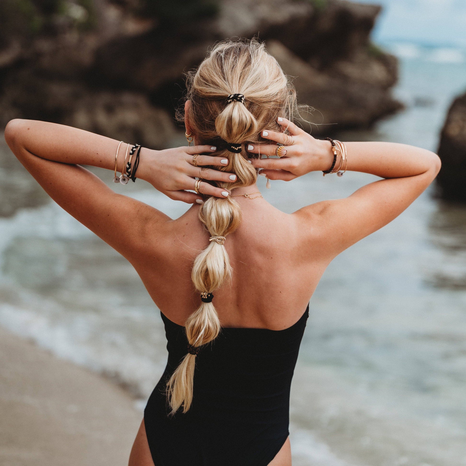 Woman in a black swimsuit with braided hair standing on a beach with rocky cliffs and ocean in the background.