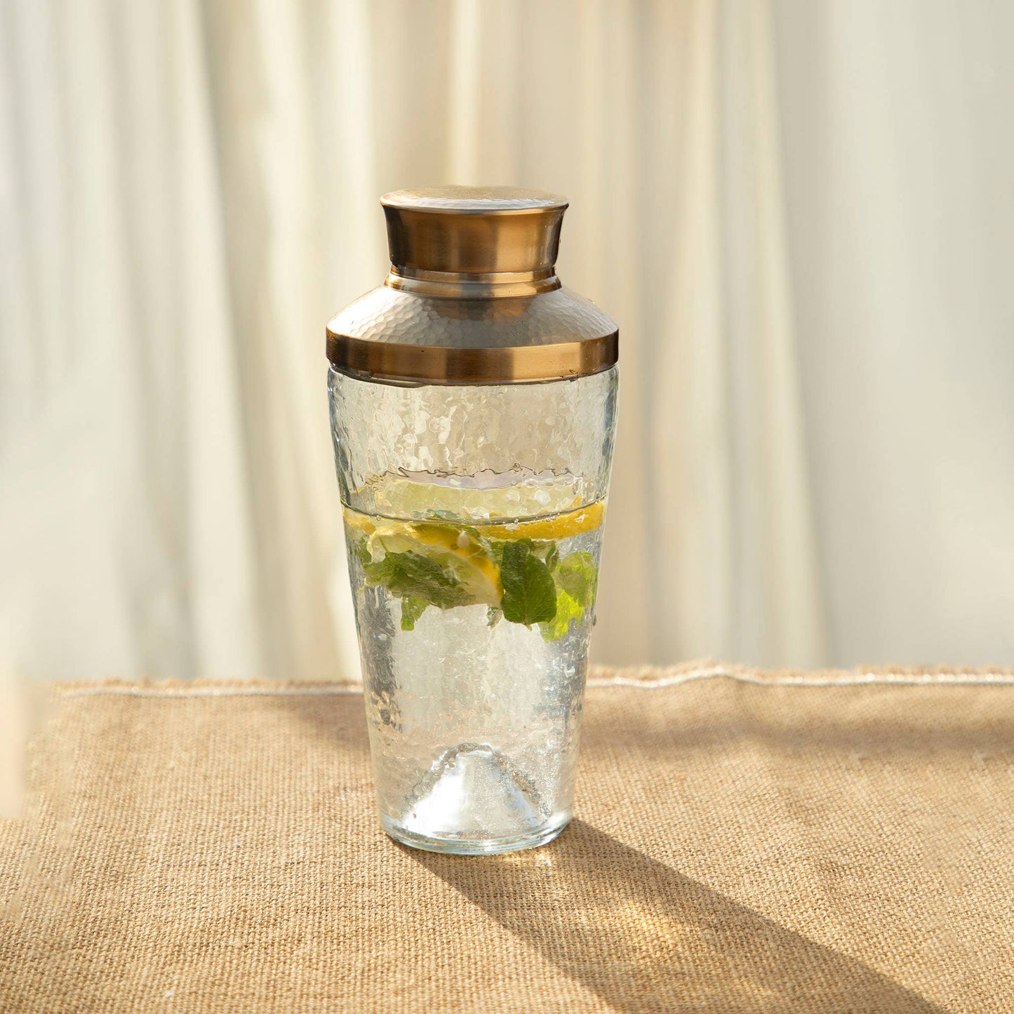 Clear glass carafe with gold lid containing water and lemon slices on a beige surface.