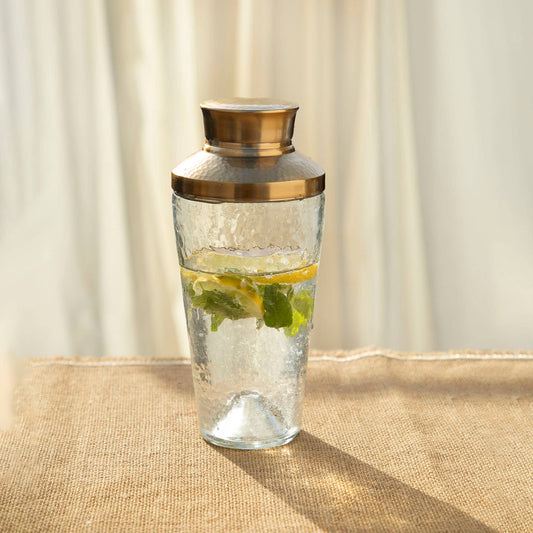 Clear glass carafe with gold lid containing water and lemon slices on a beige surface.